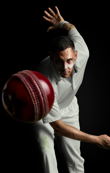 Cricketer Scott Boland bowls a ball directly at the camera. The ball appears at close range suspended in mid-air. Scott leans forwards with one arm outstretched behind him, indicating an action shot.