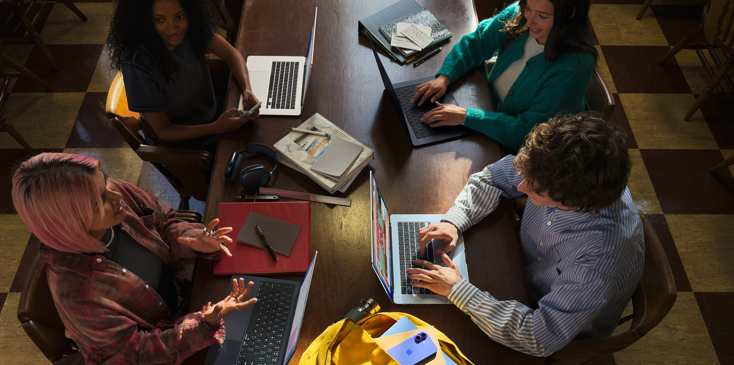 Cuatro estudiantes en una mesa. Tienen sus respectivos MacBook y apuntes abiertos frente a ellos.