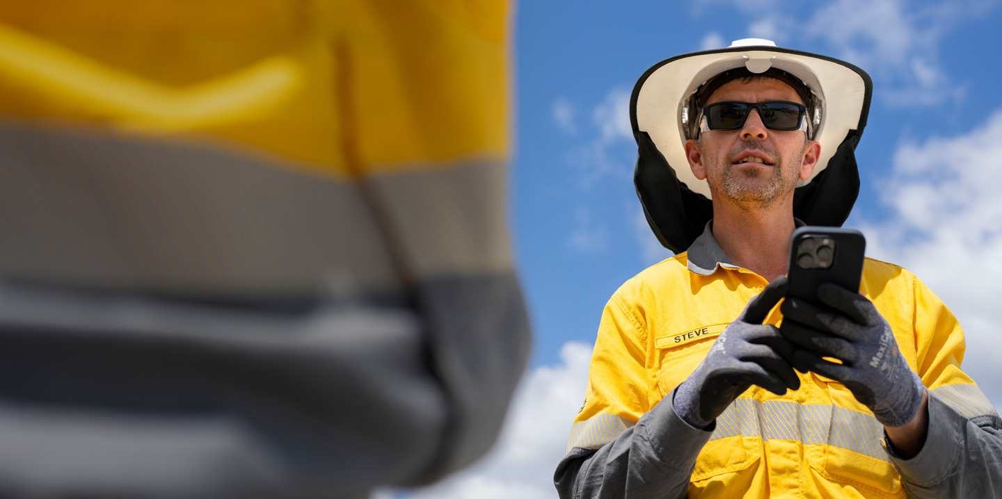 A field worker dressed in safety gear holds an iPhone while talking with a crew member