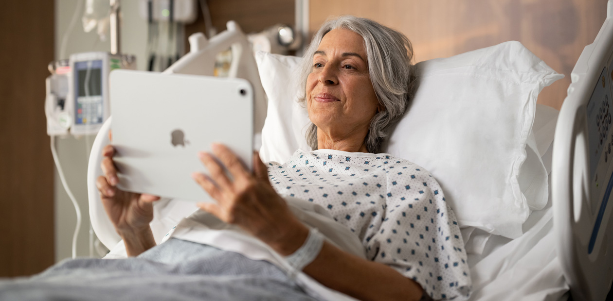 A female patient lays in hospital bed while looking at her iPad.