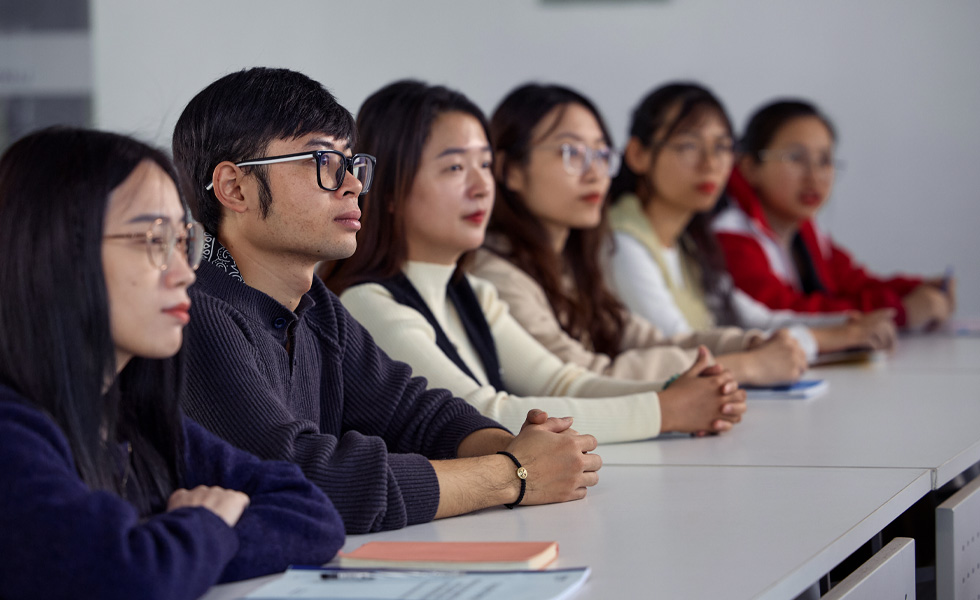 Six people sit at a long row of desks focused on the front of the classroom